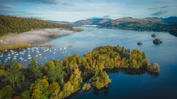 Aerial view of a serene lake surrounded by lush green and autumn-colored trees, with boats anchored near the shoreline and mist hovering above the water.
