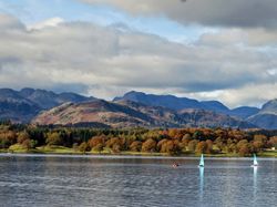 Scenic landscape with a lake, sailboats, autumn-colored trees, and mountains under a cloudy sky.