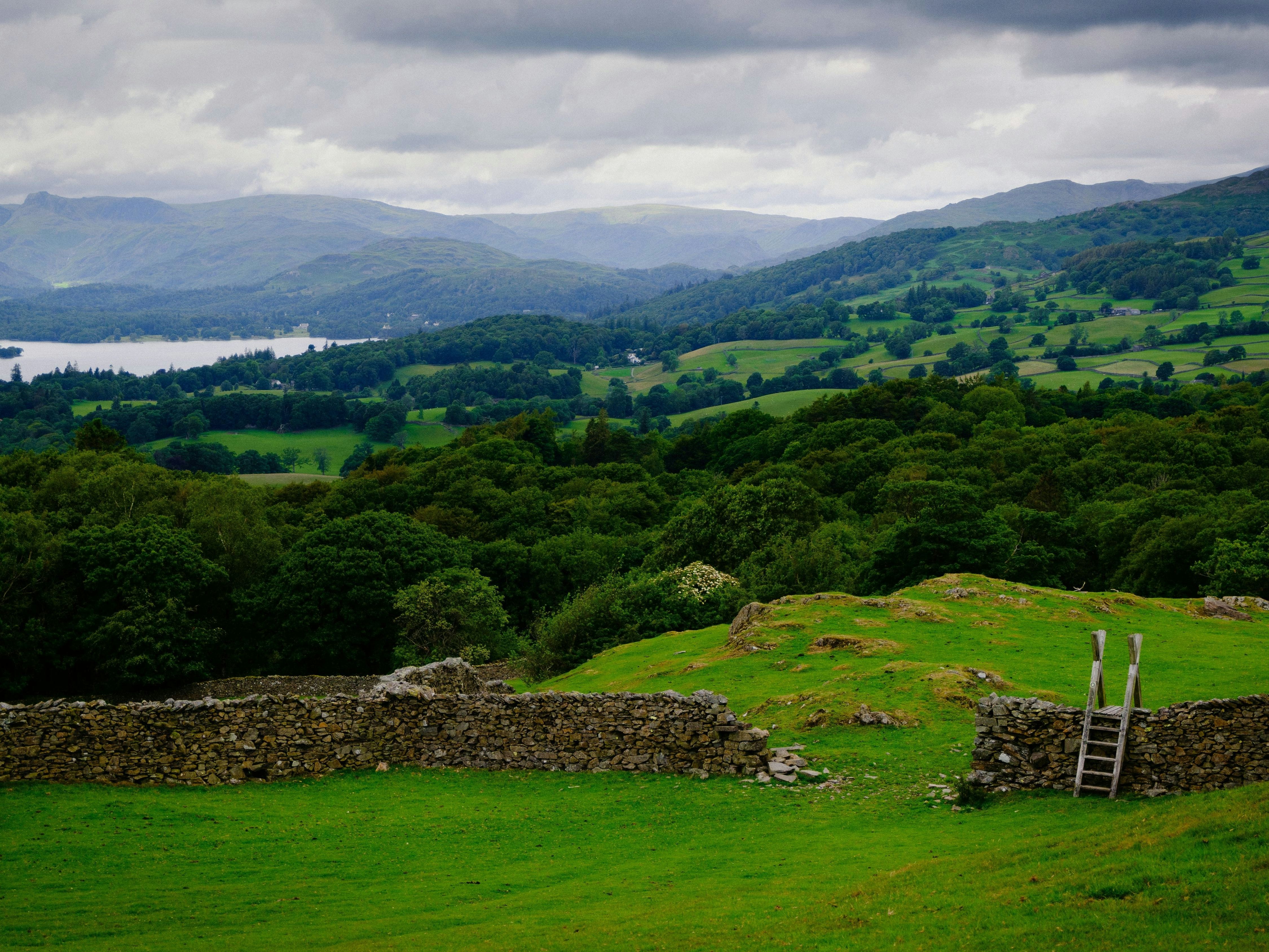 Scenic countryside landscape with green rolling hills, a stone wall with a wooden stile, and a distant lake under a cloudy sky.