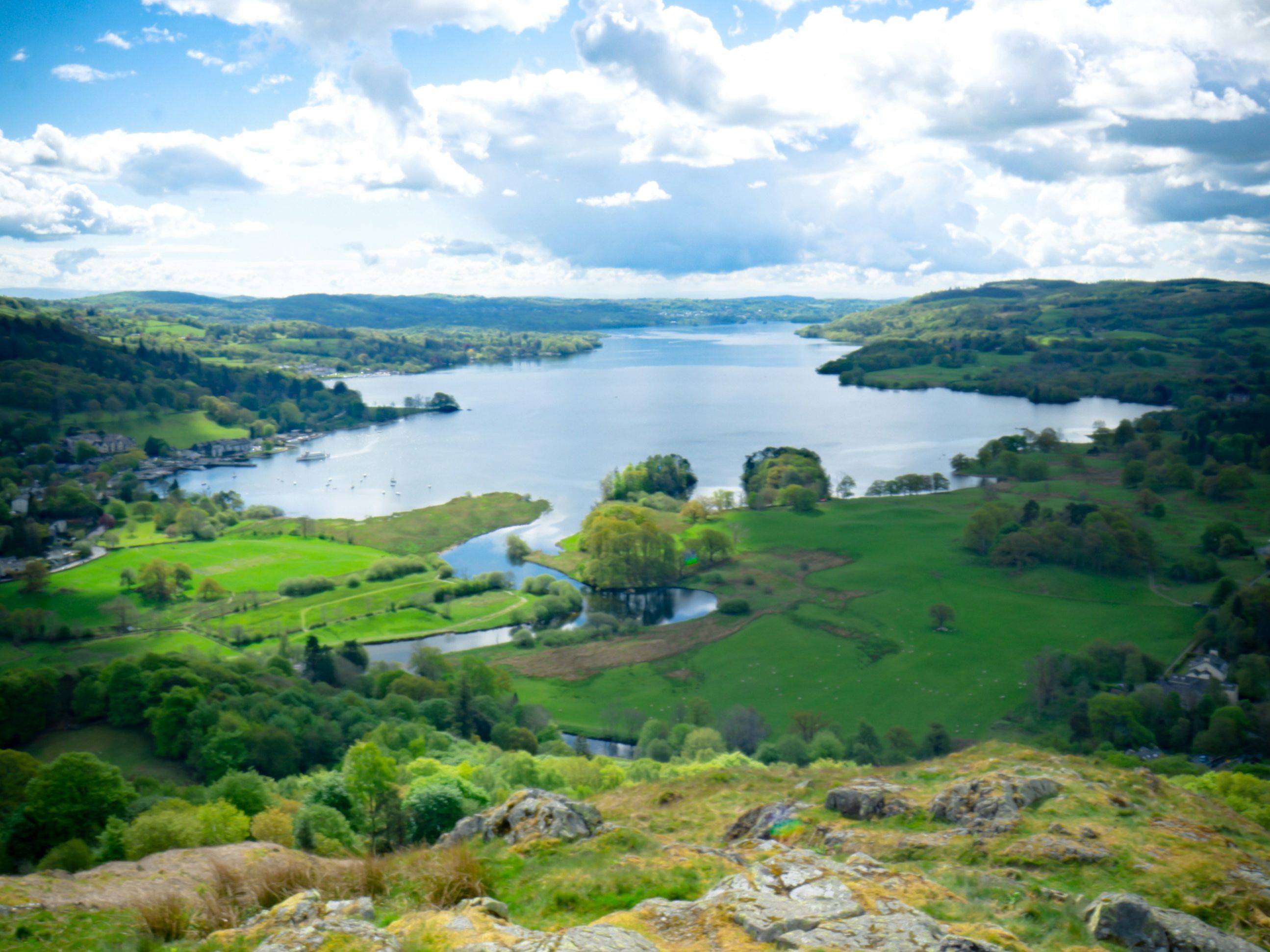 A scenic landscape view of a lake surrounded by green hills and rocky terrain under a dramatic cloudy sky.