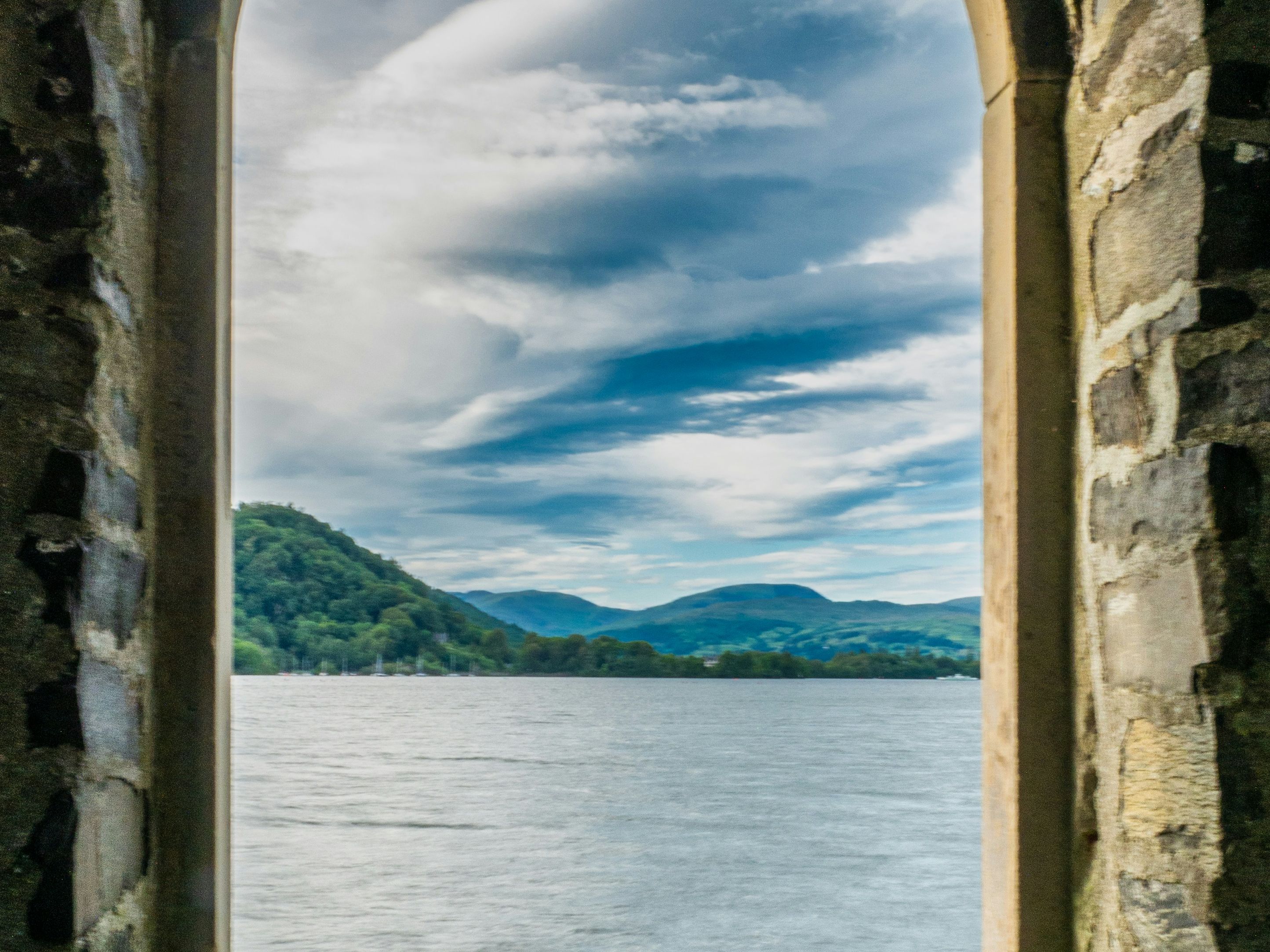 Stone arch window overlooking a lake and green hills under a cloudy blue sky