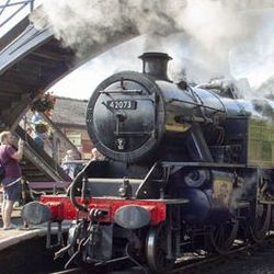 Steam locomotive train at a station with people taking photos and a footbridge in the background.