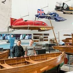 Two people standing inside a wooden boat at a museum with several historical boats and speedboats displayed around and above them.