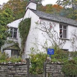 White stone cottage with ivy on its walls, surrounded by greenery and stone fence