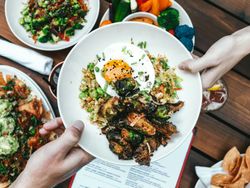 People sharing a bowl of food with roasted vegetables, fried egg, and rice on a table with other plates of food.