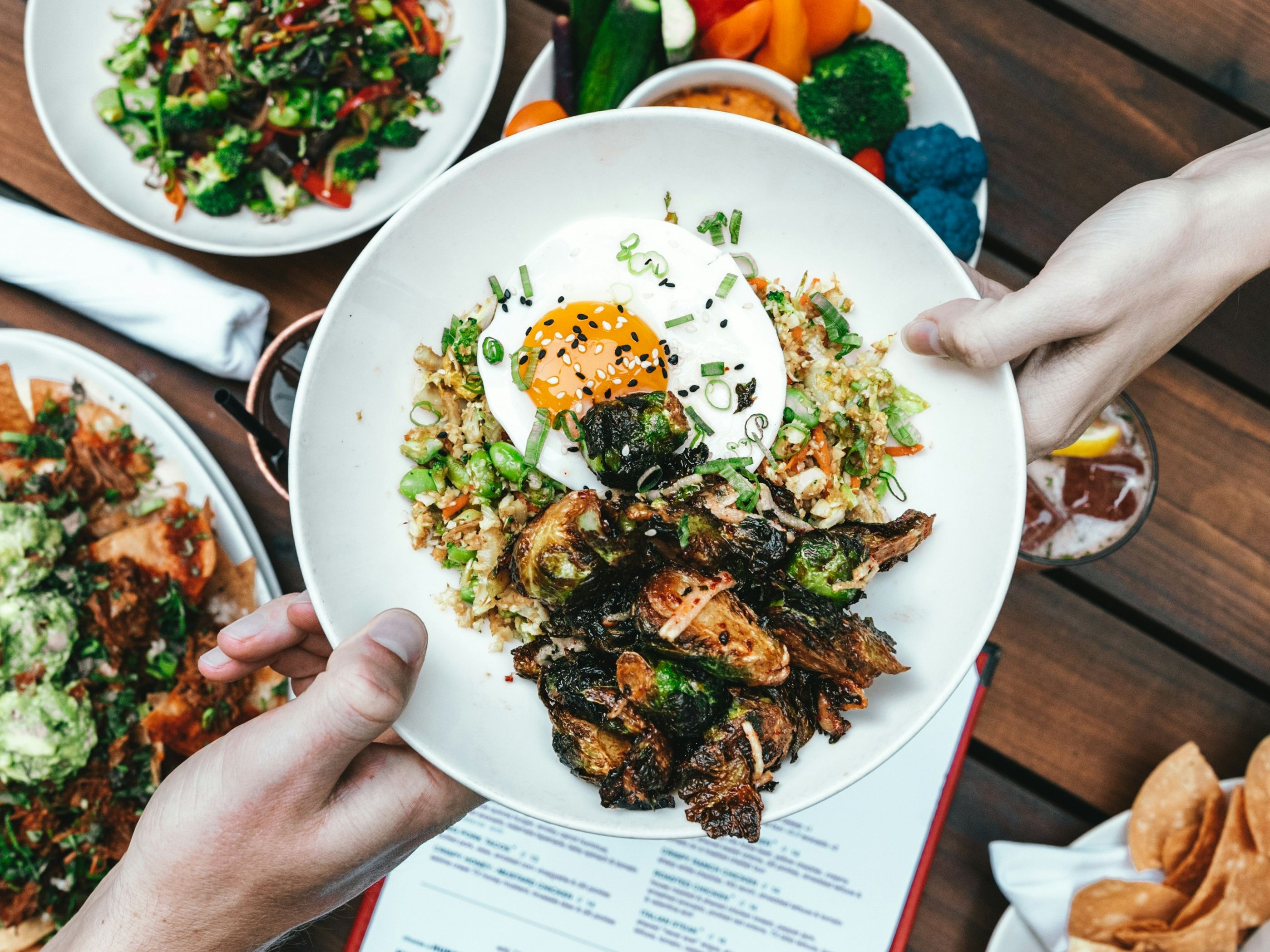 People sharing a bowl of food with roasted vegetables, fried egg, and rice on a table with other plates of food.
