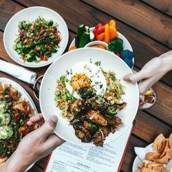 People sharing a bowl of food with roasted vegetables, fried egg, and rice on a table with other plates of food.