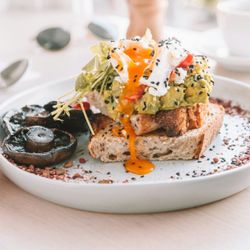 Sourdough toast topped with avocado, poached egg, feta, seeds, greens, and served with grilled mushrooms.