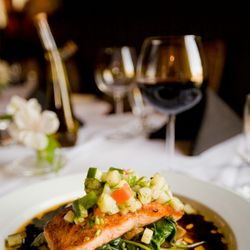 Gourmet salmon fillet topped with fresh vegetable salsa on a white plate, served with sauce and greens in a fine dining setting.
