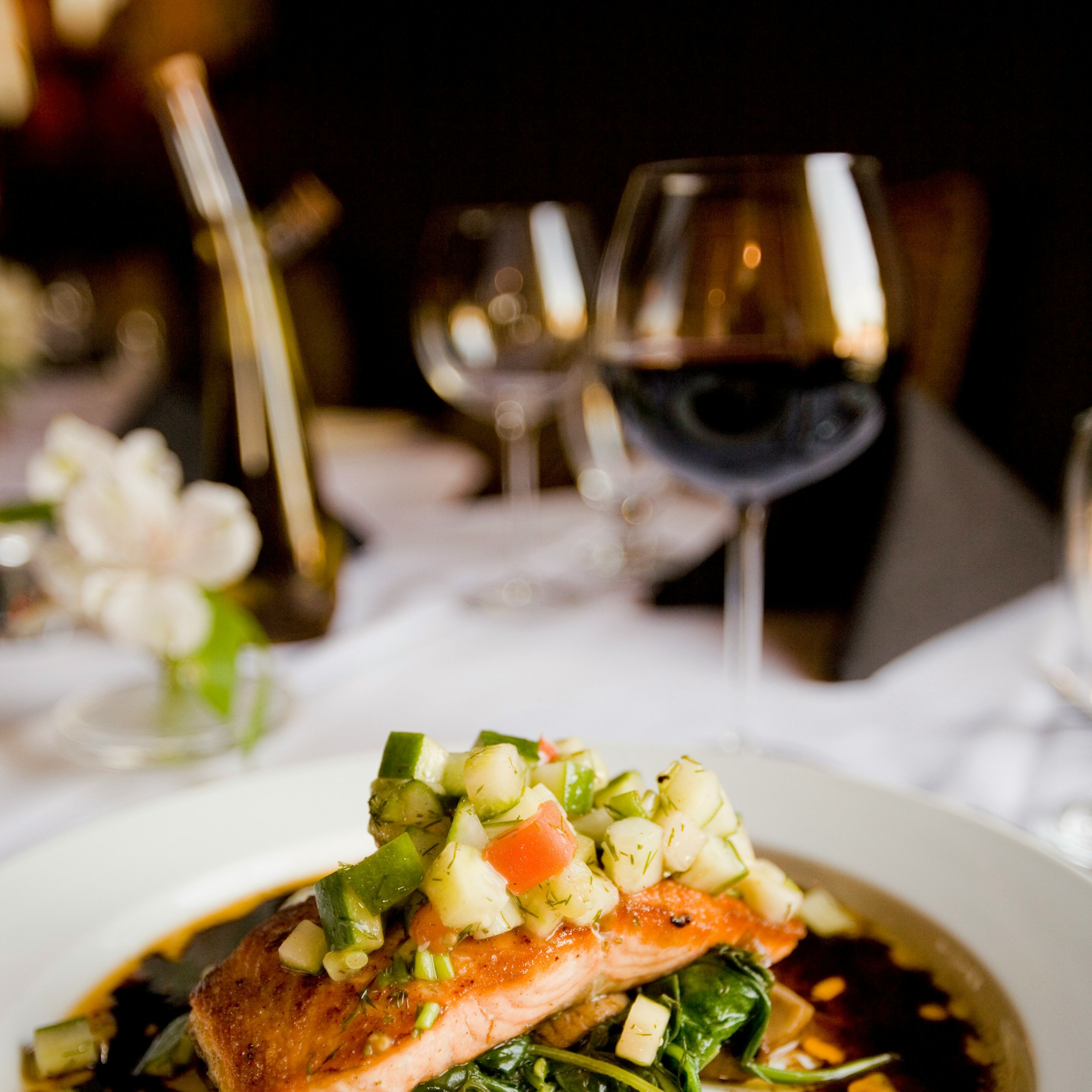 Gourmet salmon fillet topped with fresh vegetable salsa on a white plate, served with sauce and greens in a fine dining setting.