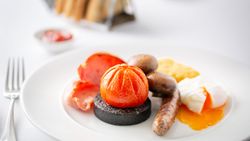 Full English breakfast with sausage, black pudding, grilled tomato, mushrooms, bacon, hash brown, and poached egg on a white plate.