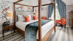 Luxurious bedroom with a four-poster bed, colorful bird-themed wallpaper, two orange chairs, and blue curtains.