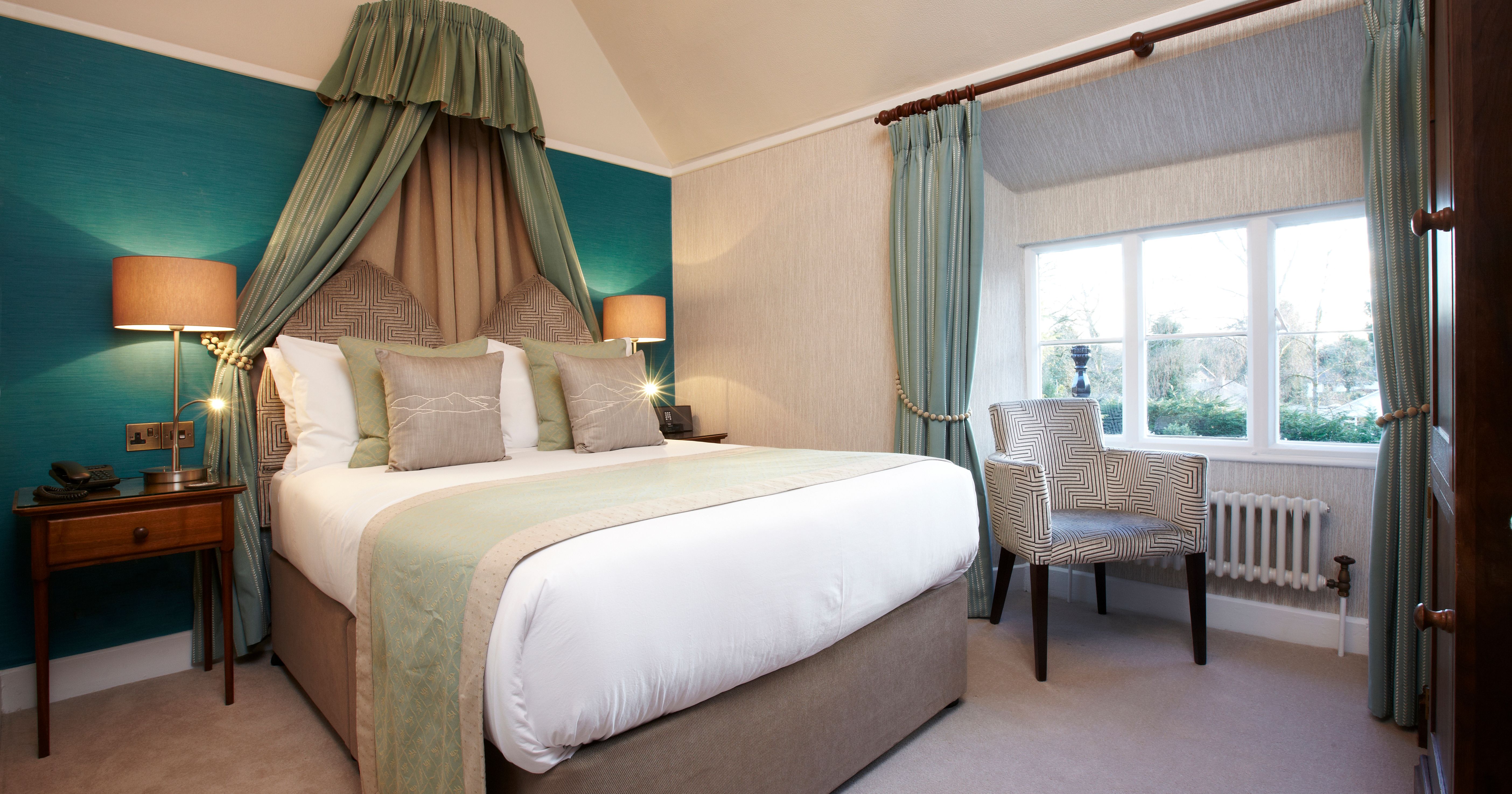 Elegant bedroom with a double bed, teal and beige decor, bedside lamps, armchair, and a large window with blue curtains.