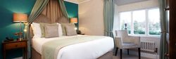 Elegant bedroom with a double bed, teal and beige decor, bedside lamps, armchair, and a large window with blue curtains.