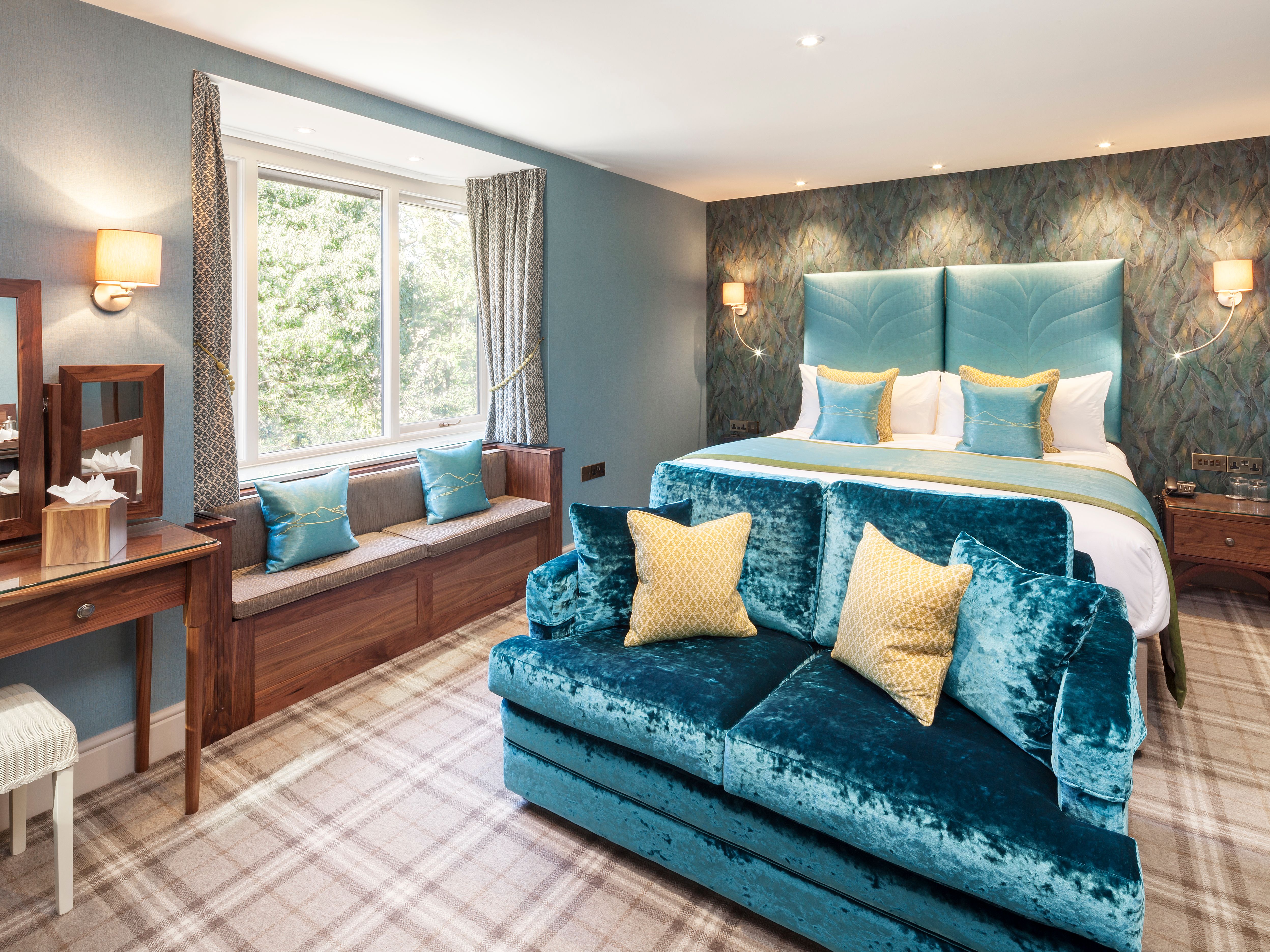 Elegant hotel room with blue and yellow accents, a large bed, sofa, vanity, and window seat.