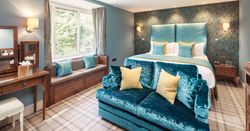 Elegant hotel room with blue and yellow accents, a large bed, sofa, vanity, and window seat.