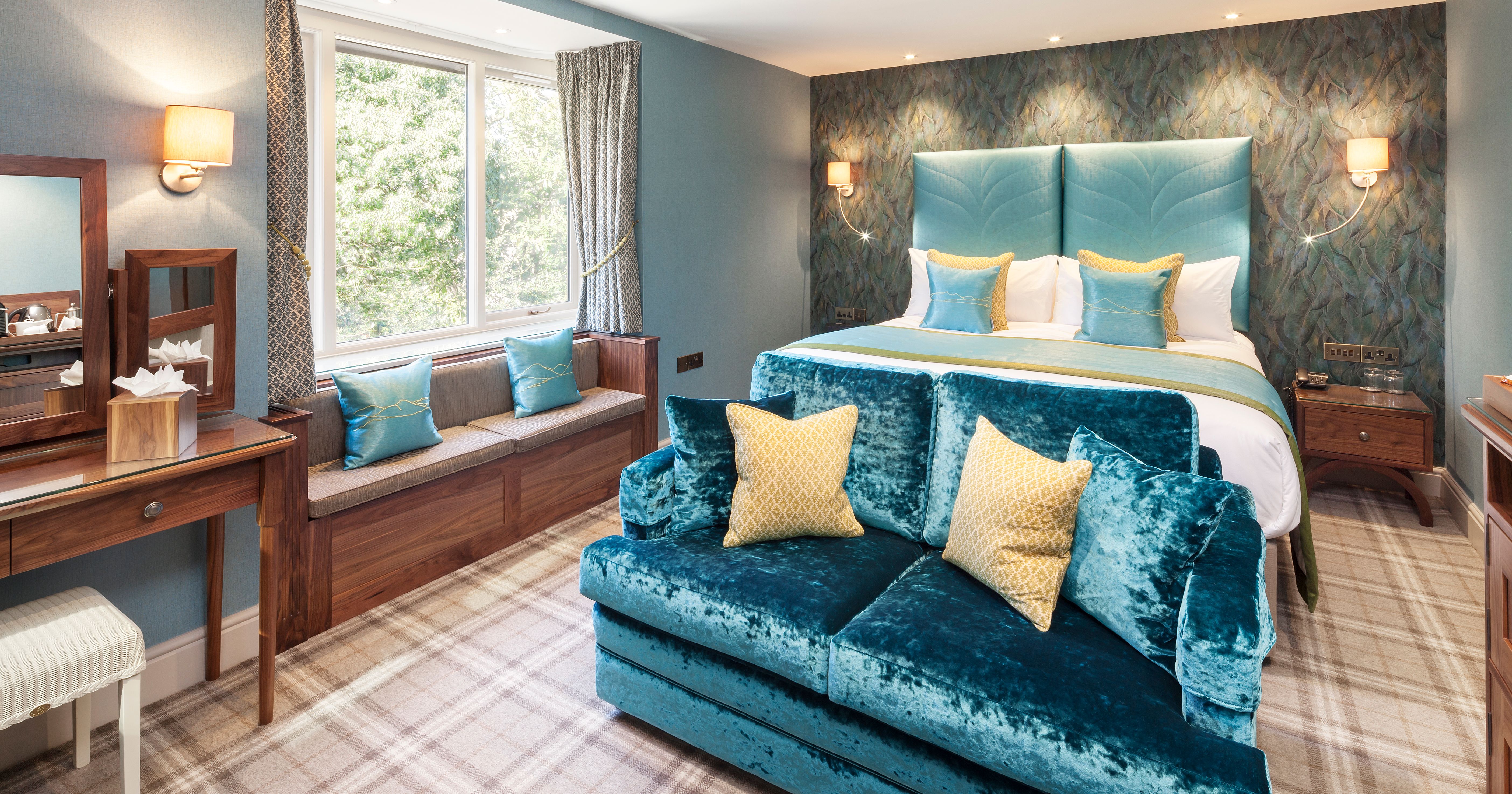 Elegant hotel room with blue and yellow accents, a large bed, sofa, vanity, and window seat.