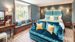 Elegant hotel room with blue and yellow accents, a large bed, sofa, vanity, and window seat.