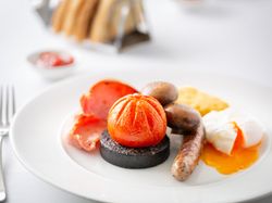 Full English breakfast with sausage, black pudding, tomato, mushrooms, bacon, hash brown, and poached egg on a white plate.