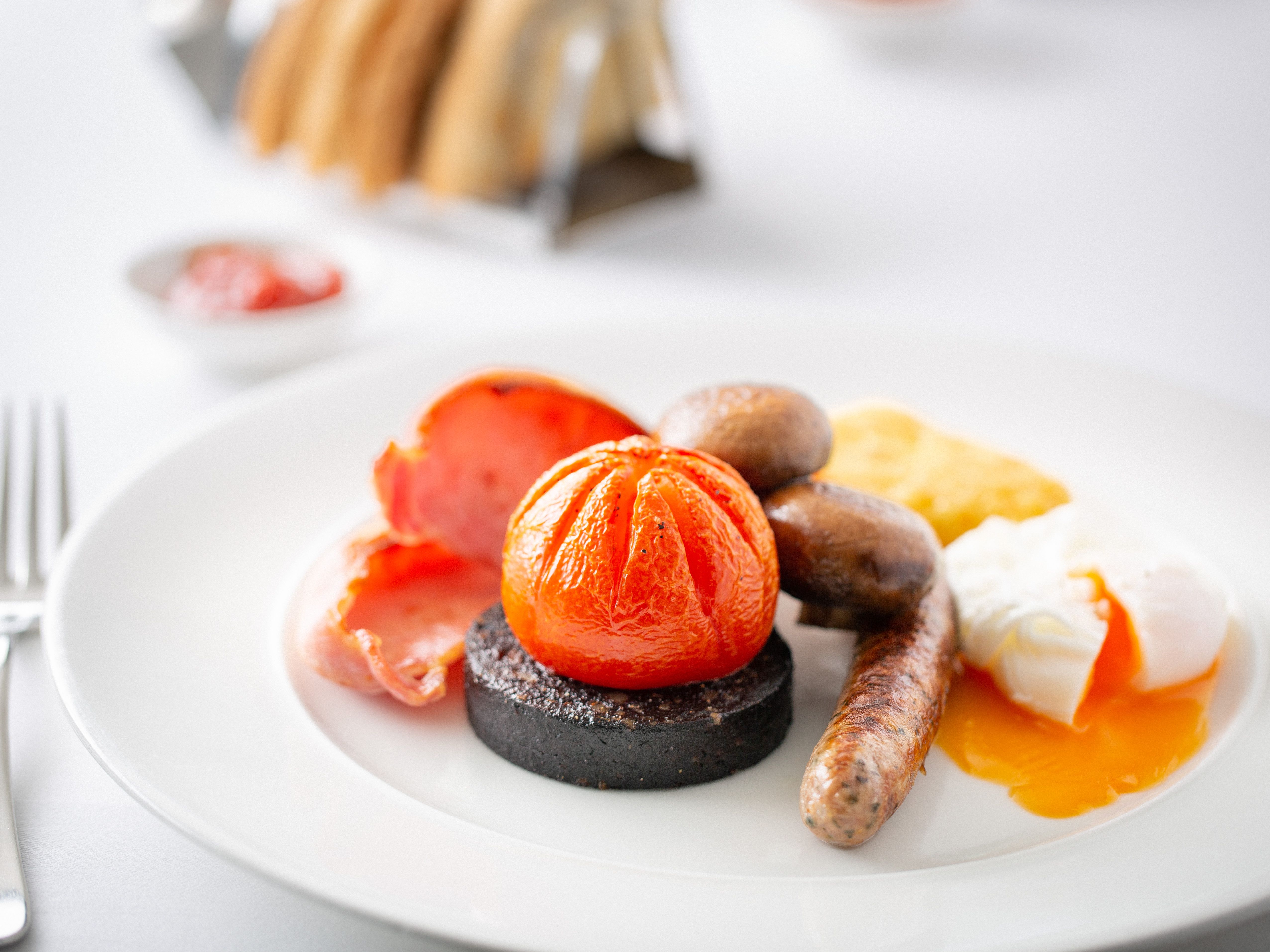 Full English breakfast with sausage, black pudding, tomato, mushrooms, bacon, hash brown, and poached egg on a white plate.