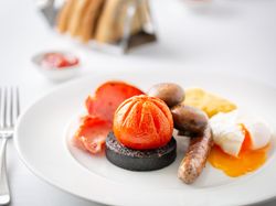 Full English breakfast with sausage, black pudding, tomato, mushrooms, bacon, hash brown, and poached egg on a white plate.