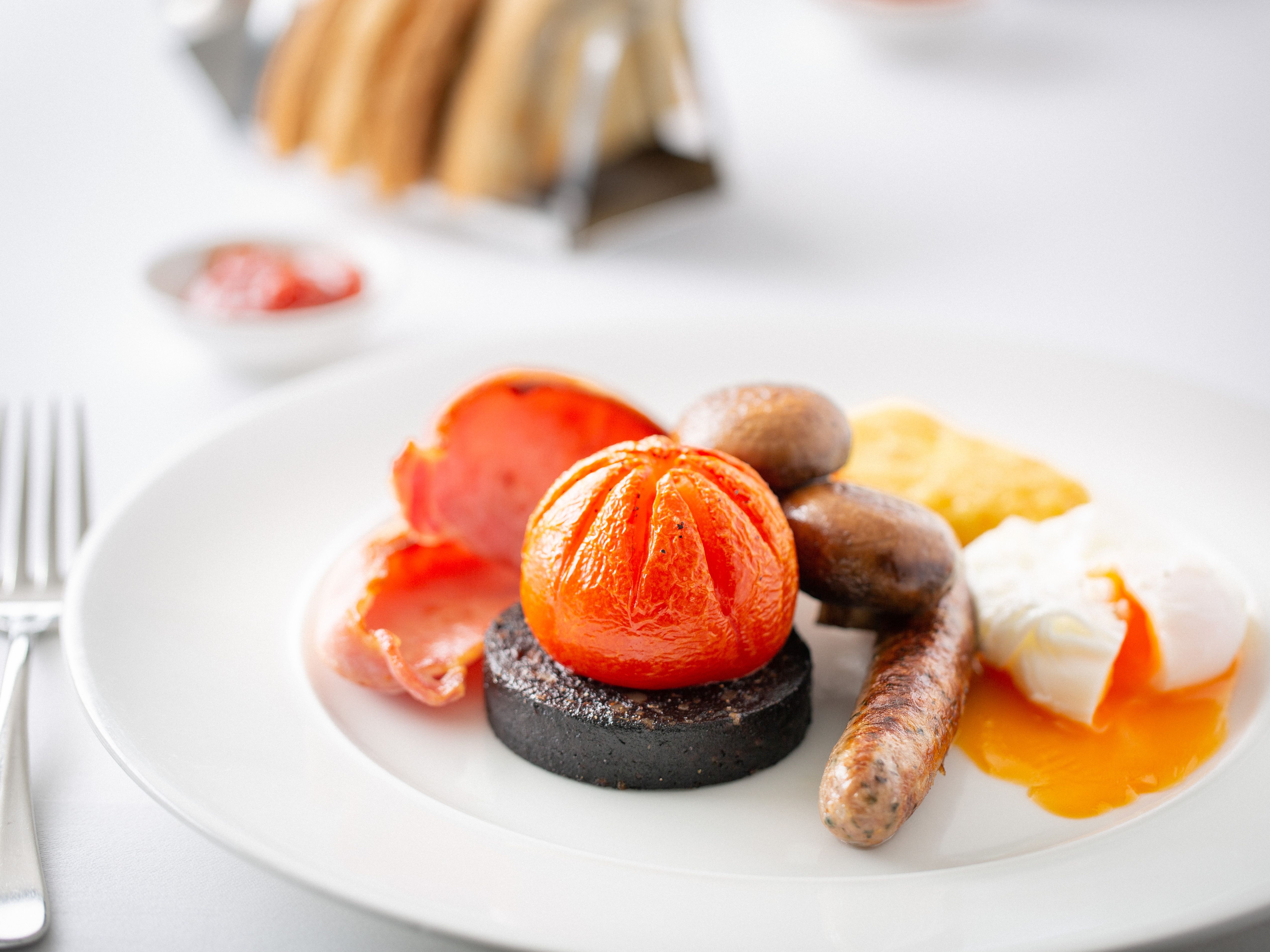 Full English breakfast with sausage, black pudding, tomato, mushrooms, bacon, hash brown, and poached egg on a white plate.