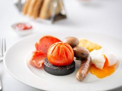 Full English breakfast with sausage, black pudding, tomato, mushrooms, bacon, hash brown, and poached egg on a white plate.