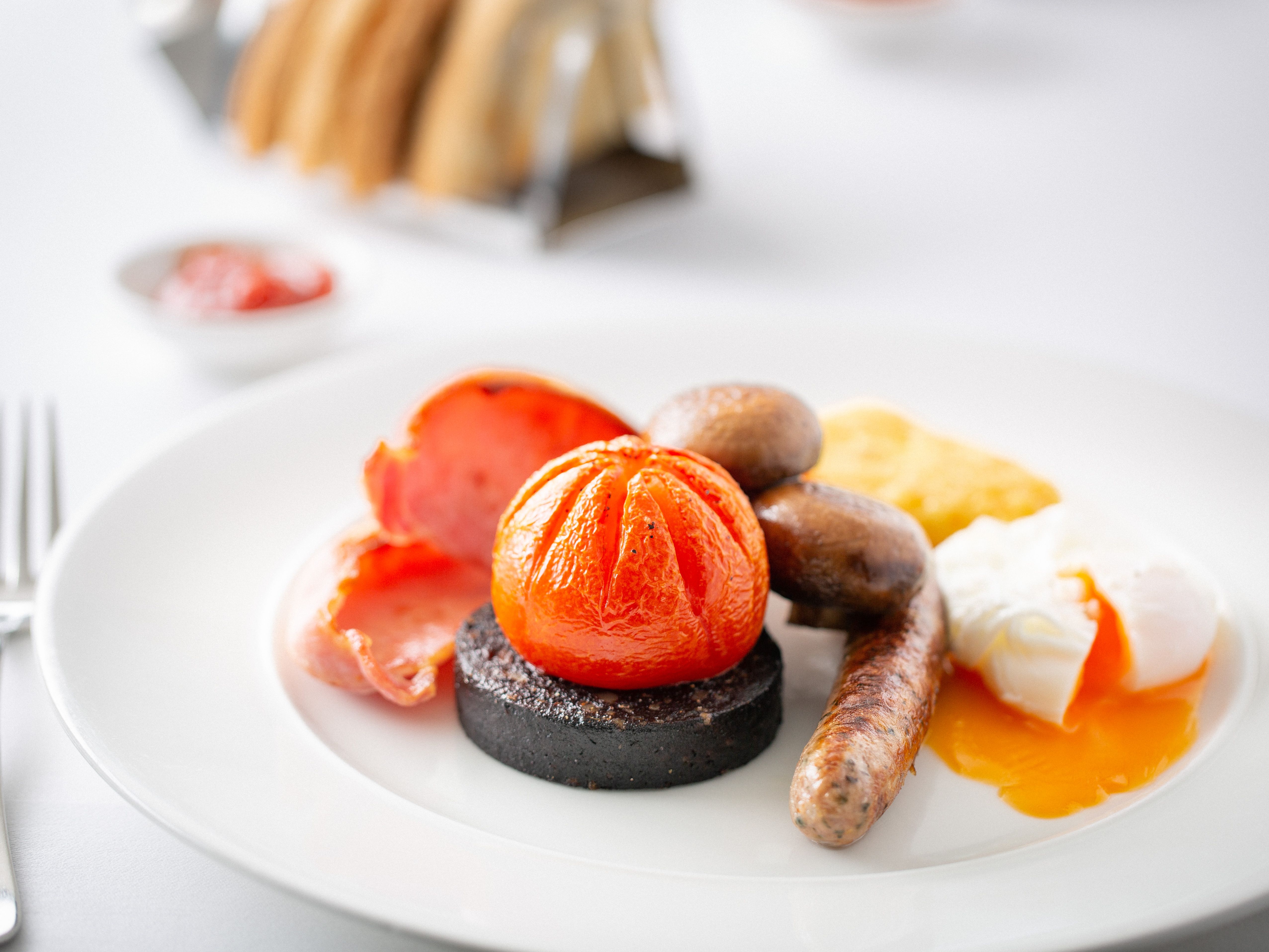 Full English breakfast with sausage, black pudding, tomato, mushrooms, bacon, hash brown, and poached egg on a white plate.