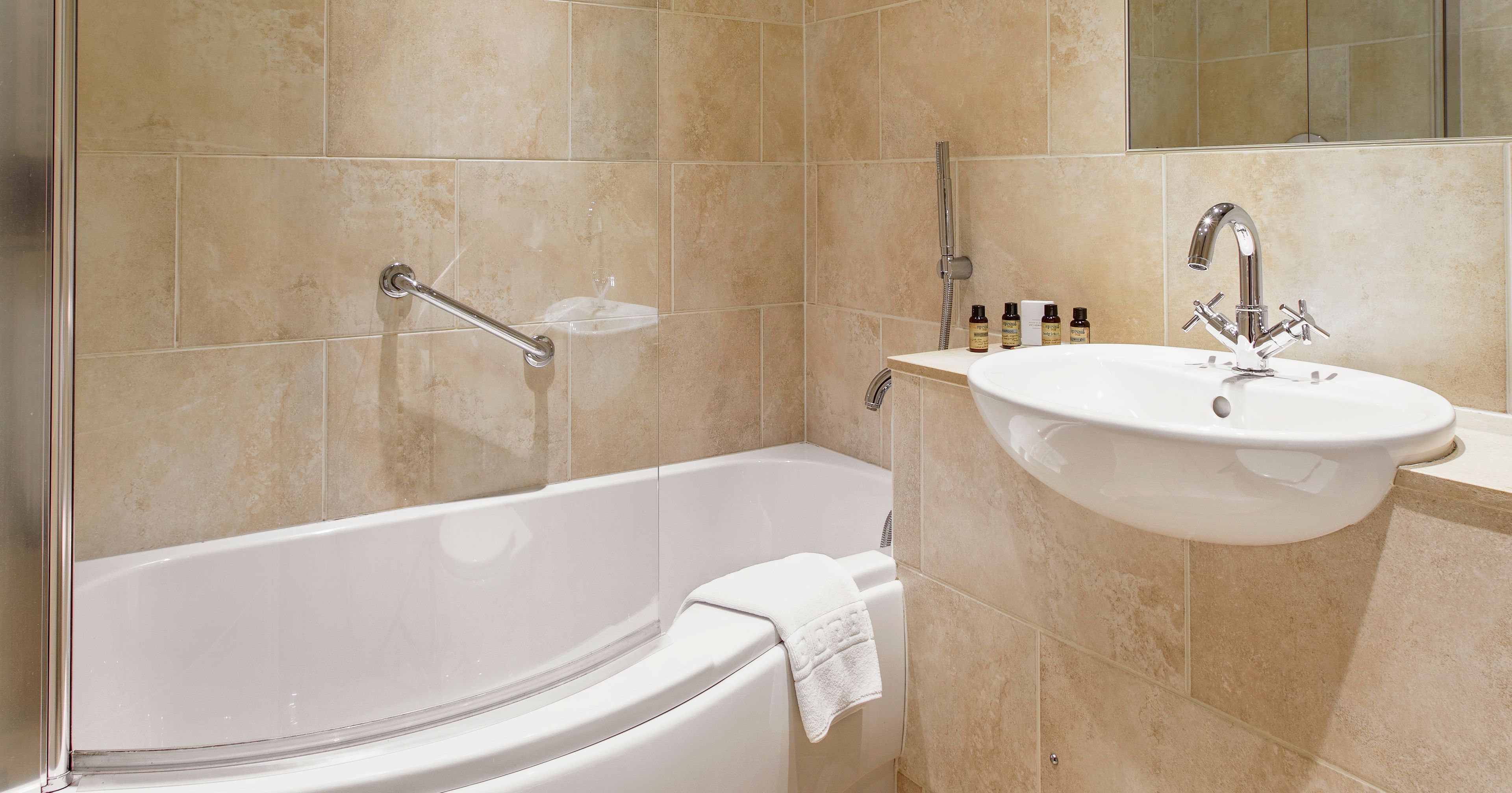 Modern bathroom with tan tile, curved bathtub, glass shower screen, wall-mounted sink, mirror, and toiletries.