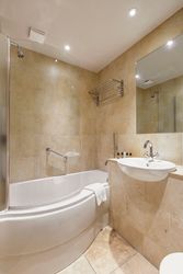 Modern bathroom with tan tile, curved bathtub, glass shower screen, wall-mounted sink, mirror, and toiletries.