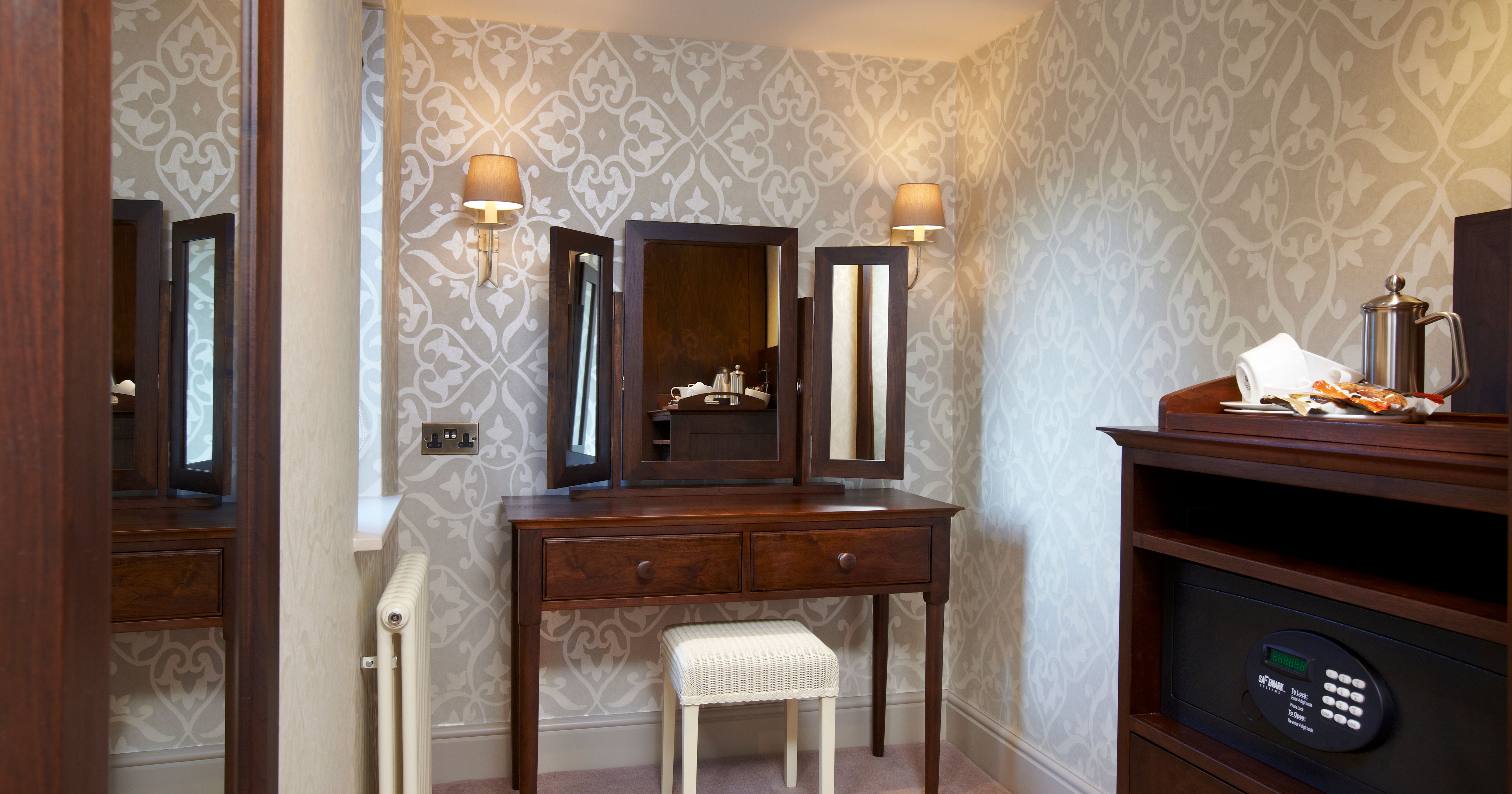 Elegant dressing table with mirror, wooden drawers, and a cushioned stool in a patterned wallpaper room.