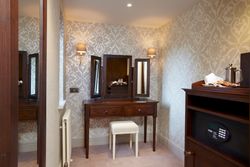 Elegant dressing table with mirror, wooden drawers, and a cushioned stool in a patterned wallpaper room.