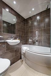 Modern bathroom with brown tiled walls, a white bathtub with glass shower screen, a wall-mounted sink, and toilet.