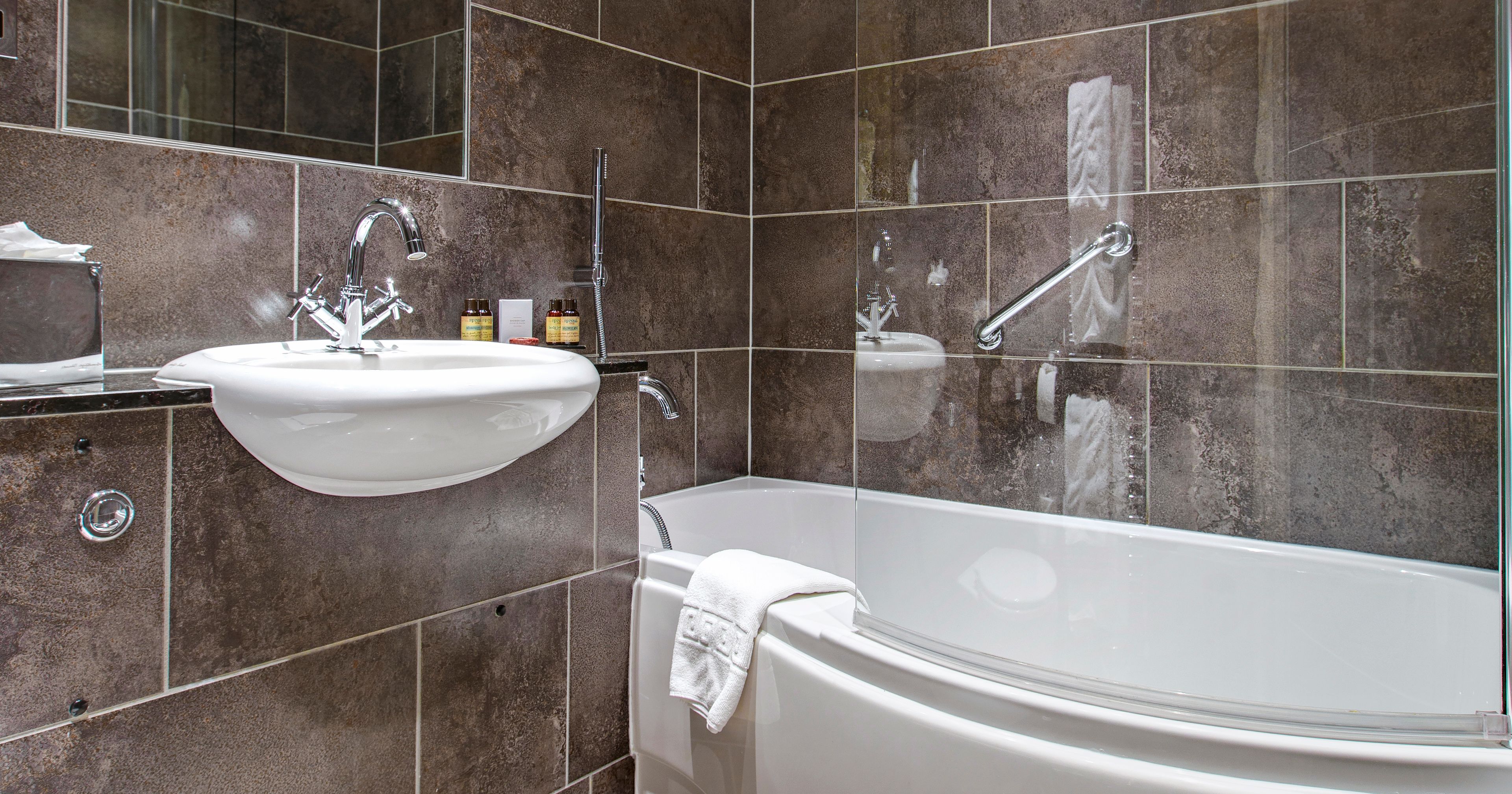 Modern bathroom with brown tiled walls, a white bathtub with glass shower screen, a wall-mounted sink, and toilet.