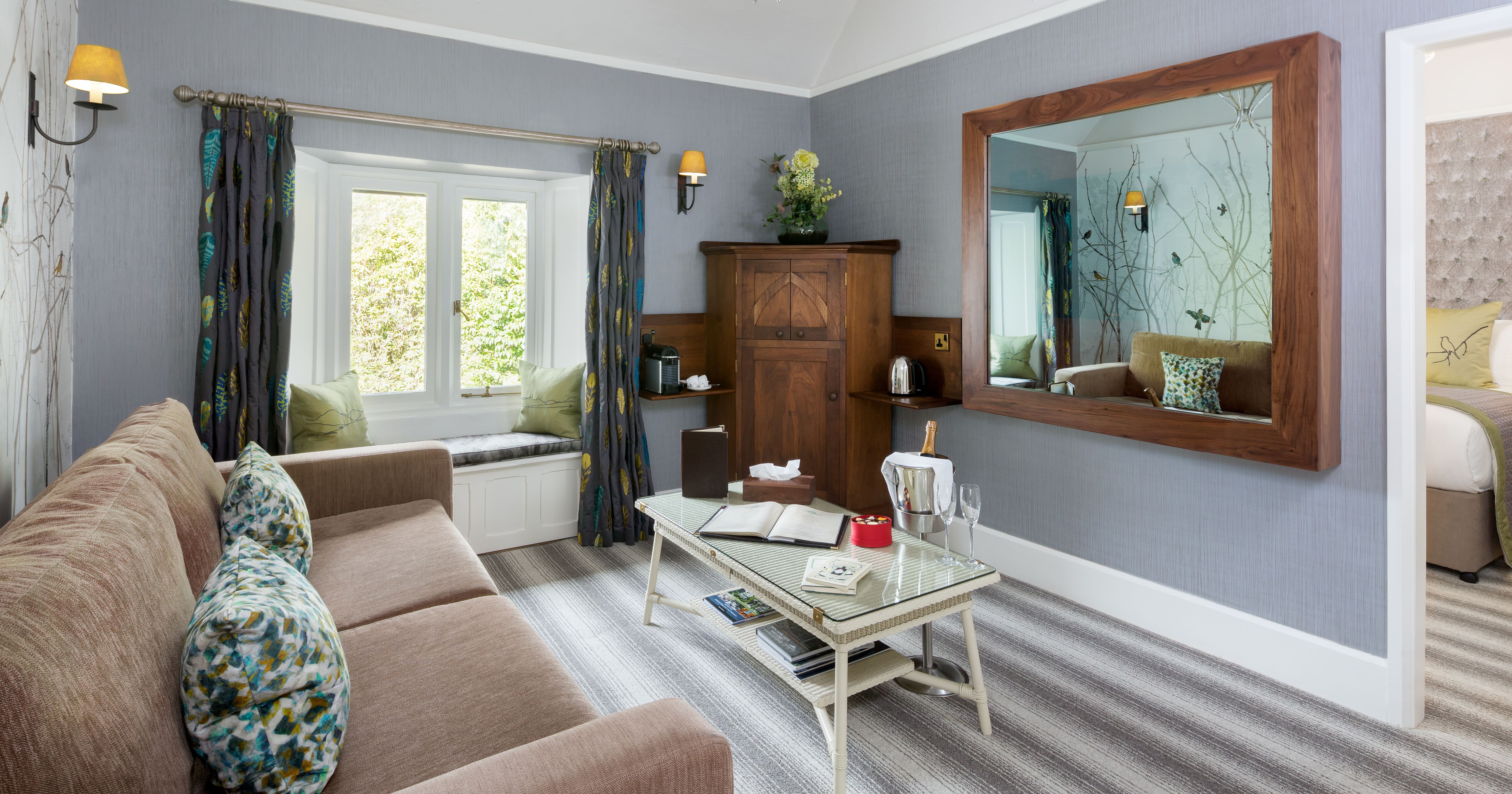 Modern lounge area with beige sofa, large mirror, and window seat in a hotel suite.