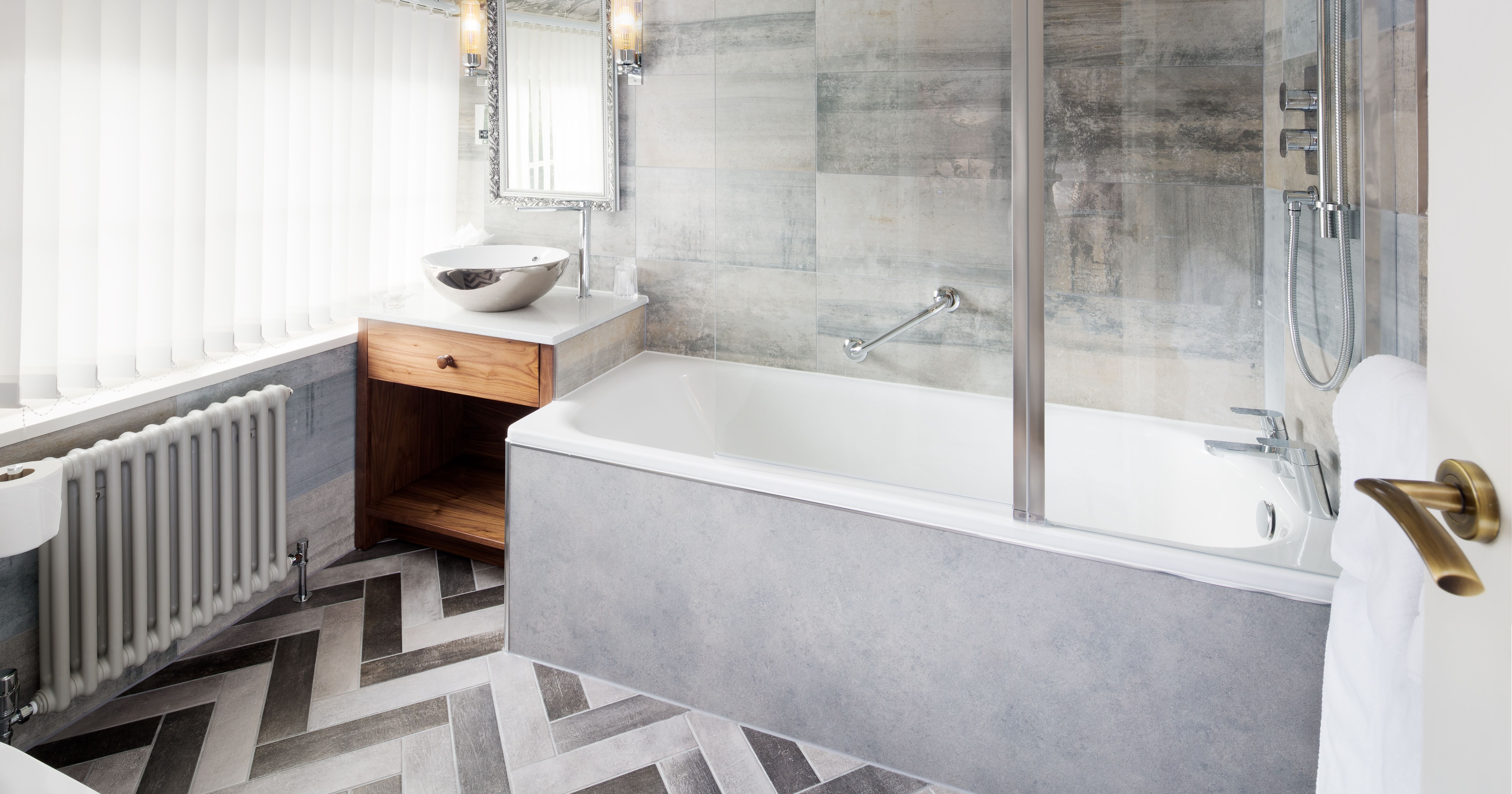 Modern en-suite bathroom with bathtub, patterned flooring, chandelier, and large window with vertical blinds.