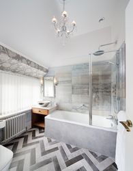 Modern en-suite bathroom with bathtub, patterned flooring, chandelier, and large window with vertical blinds.