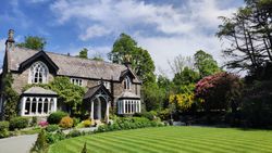 Charming stone house with arched windows surrounded by lush green garden and manicured lawn under a blue sky.