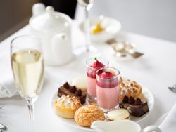 Plate of assorted desserts with glasses of mousse, macarons, brownies, and tarts on a white tablecloth, with a glass of champagne and teapot in the background.