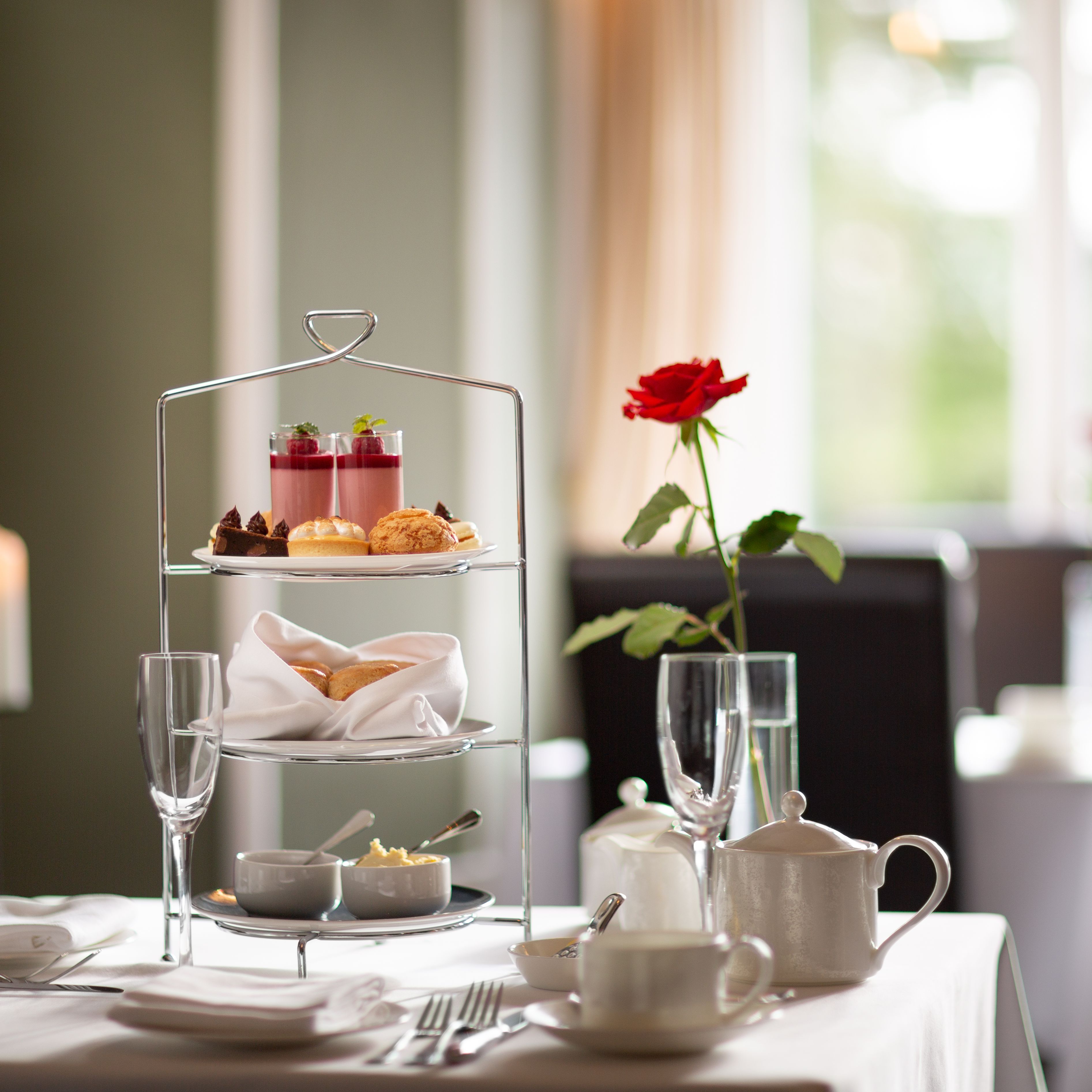 Elegant afternoon tea setup with a three-tiered stand of pastries, scones, and desserts, with a red rose in a vase on a white tablecloth.