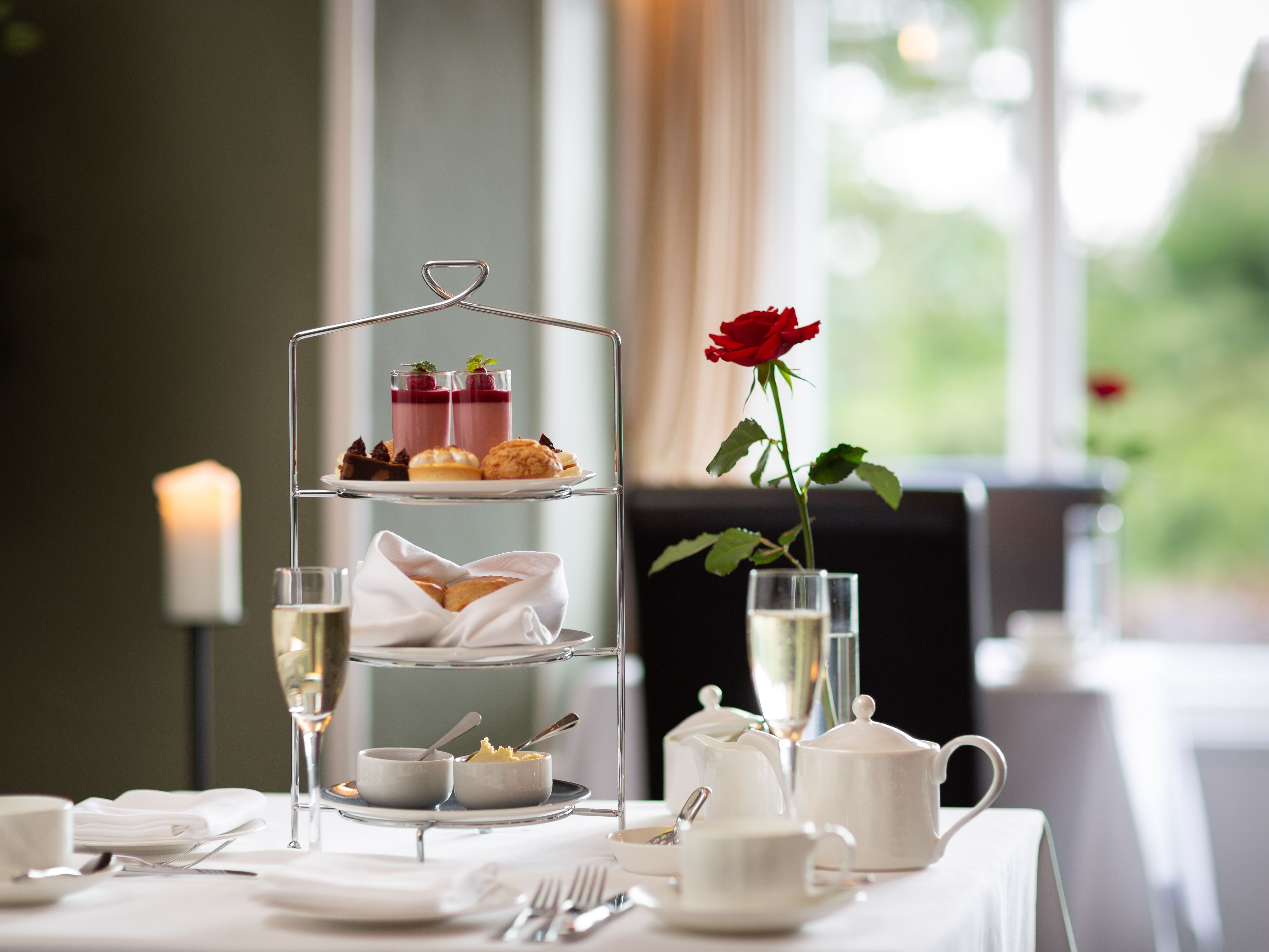Elegant afternoon tea setup with a tiered tray of pastries, a red rose in a vase, and a glass of champagne on a white tablecloth.
