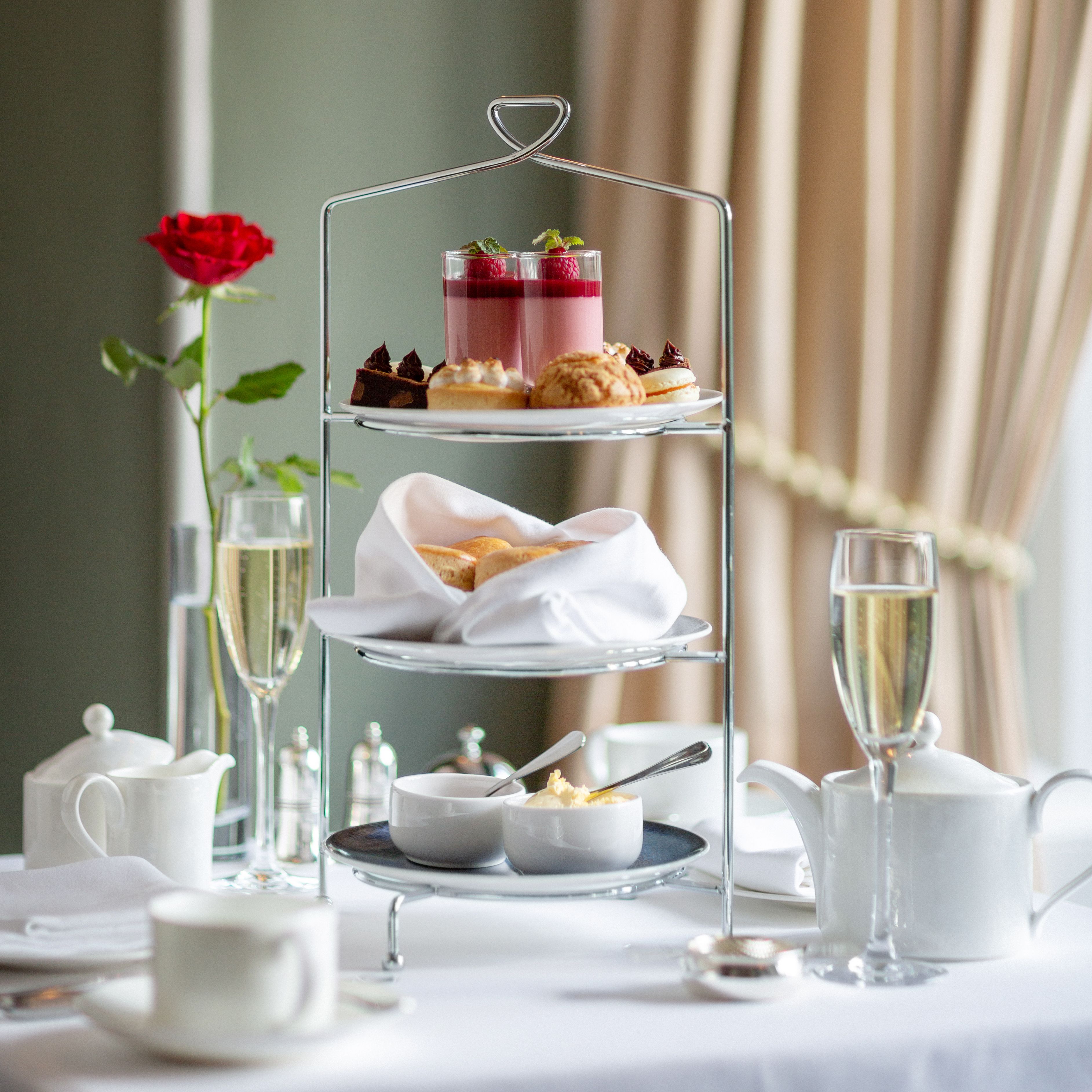 Elegant afternoon tea setup with a tiered tray of desserts and scones, tea sets, champagne, and a red rose on a white tablecloth.