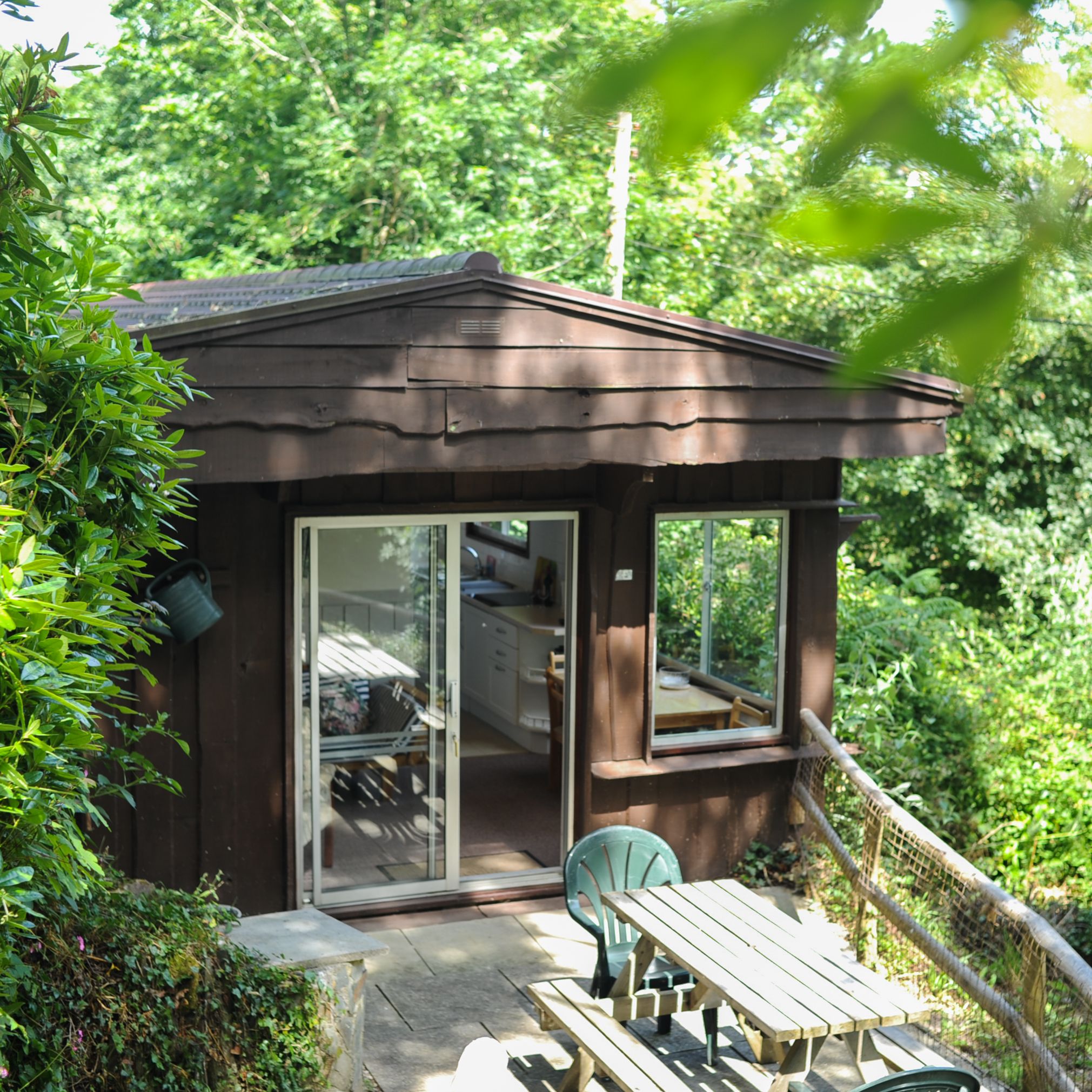 A small wooden cabin surrounded by lush greenery, with a picnic table and chairs on a patio in front.