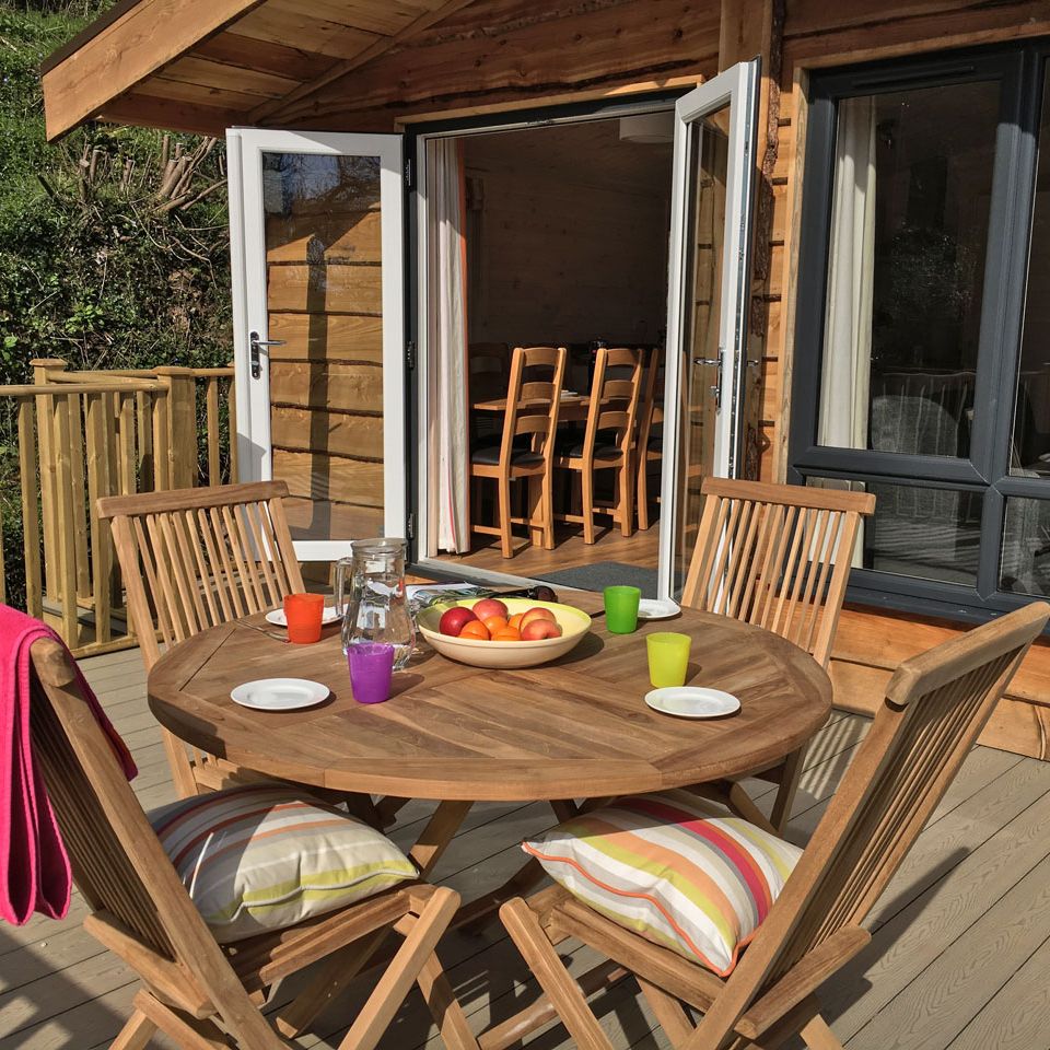 Wooden outdoor dining table with chairs on a deck, adjacent to a cabin with open doors.