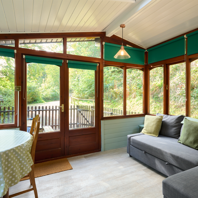 Bright sunroom with green cushions, round dining table, and large windows overlooking a lush garden.