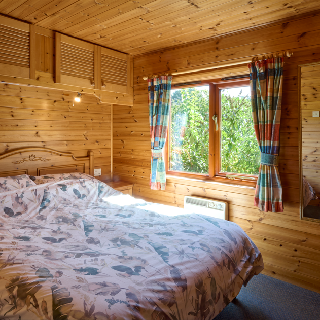 Cozy wooden bedroom with a double bed, window, colorful curtains, and a wall mirror.