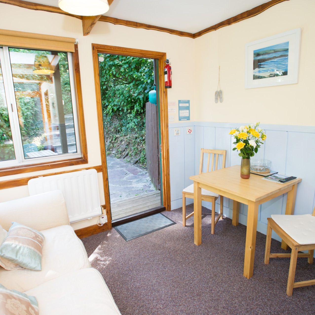 Cozy cottage interior with a small dining table, two chairs, a sofa, and an open door leading to a garden.