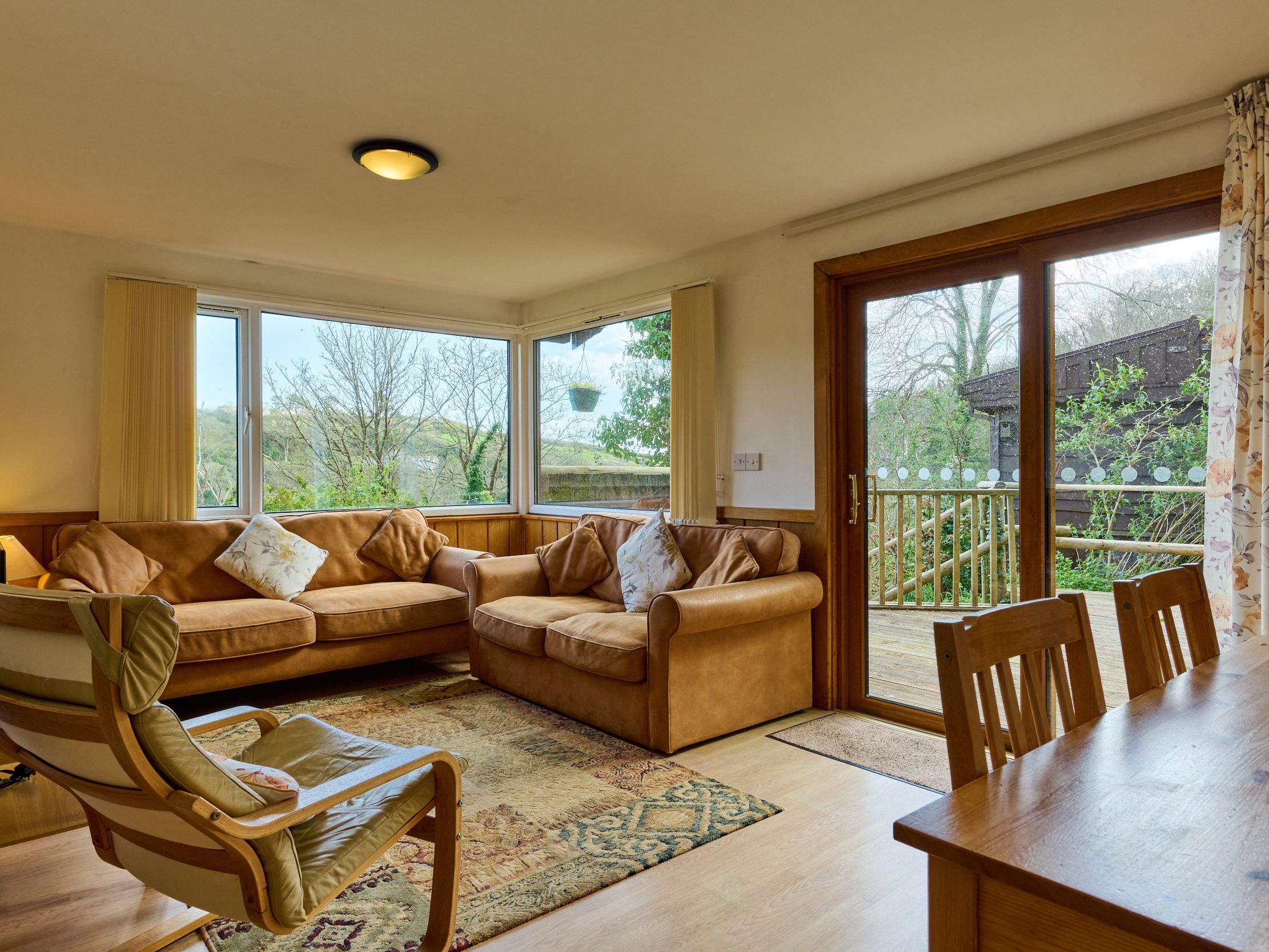 Cozy living room with tan sofas, wooden furniture, large windows, and a view of greenery outside