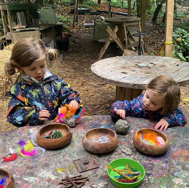 Two young children doing crafts at a paint-splattered table in a woodland outdoor classroom