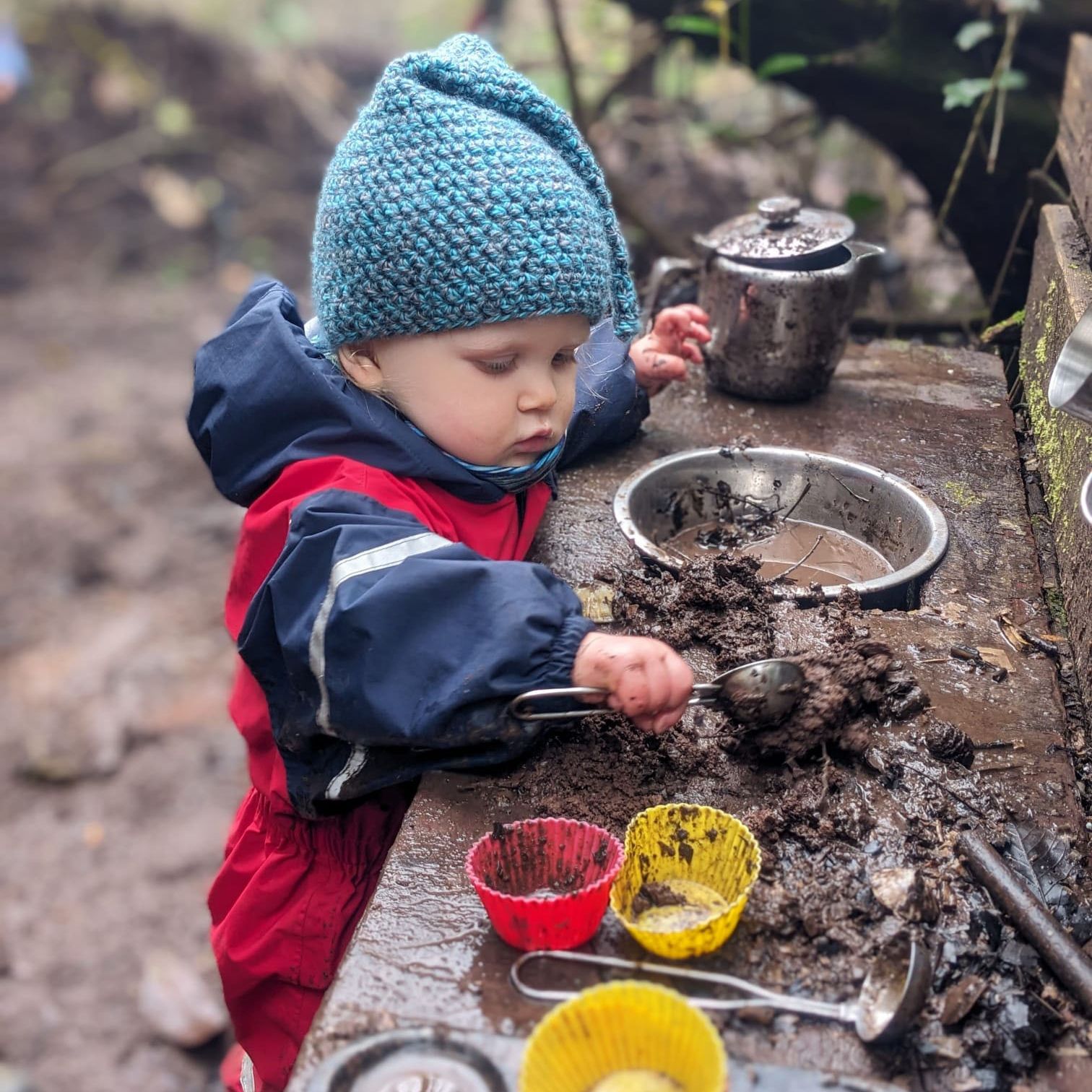 Young child in a blue knitted hat and red waterproofs playing with mud at an outdoor mud kitchen table.